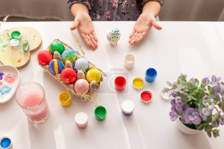Foto de "Las manos de una niña, con las palmas en alto, demuestran un huevo pintado, sobre una mesa blanca con huevos multicolores" - Imagen libre de derechos