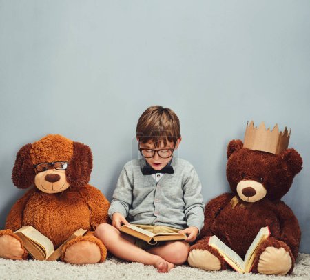 Téléchargez les photos : "Je fais des lectures. Plan studio d'un petit garçon intelligent lisant un livre à côté de ses ours en peluche sur un fond gris." - en image libre de droit