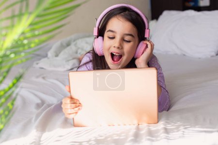 Téléchargez les photos : "Joyeux petite fille dans un casque rose couché sur un lit blanc, tenant une tablette numérique rose" - en image libre de droit