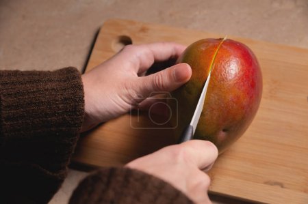 Téléchargez les photos : "Gros plan des mains de femme coupant de la mangue fraîche sur une planche à découper en bois à la cuisine maison" - en image libre de droit
