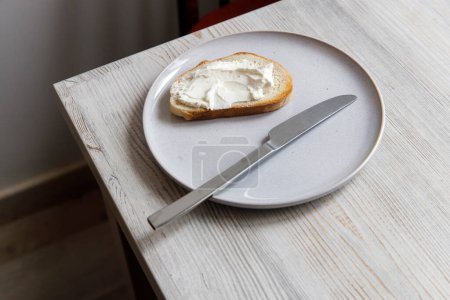 Photo for A piece of bread with curd cheese spread on it on a white ceramic plate with a knife on the beige table. - Royalty Free Image