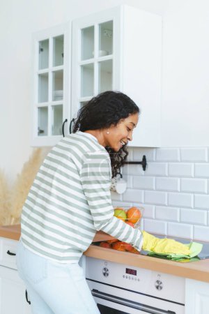 Foto de "Vista posterior de la estufa de inducción de limpieza femenina afroamericana en guantes de goma con paño" - Imagen libre de derechos