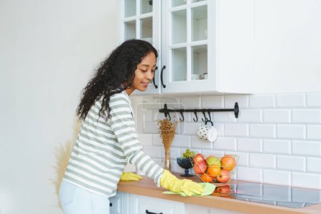 Téléchargez les photos : "Femme afro-américaine en gants de caoutchouc occupée à des tâches ménagères dans la cuisine" - en image libre de droit