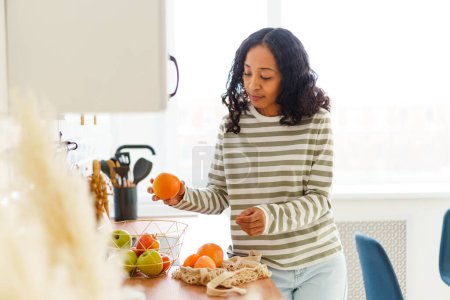 Photo for "African-American female in kitchen sorting out apples and oranges after shopping in grocery store" - Royalty Free Image