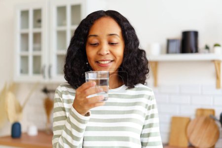 Foto de "Sonriente mujer afroamericana disfrutando de un vaso de agua clara mientras está de pie en la cocina" - Imagen libre de derechos