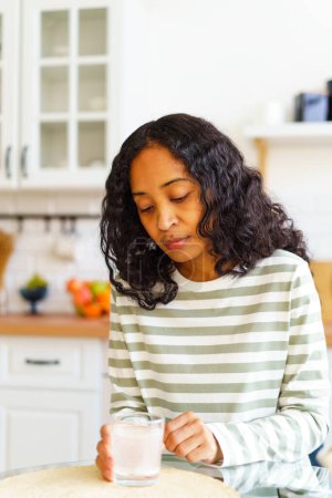 Téléchargez les photos : "Femme afro-américaine épuisée attendant la pilule de fizz pour se dissoudre dans un verre d'eau dans la cuisine" - en image libre de droit
