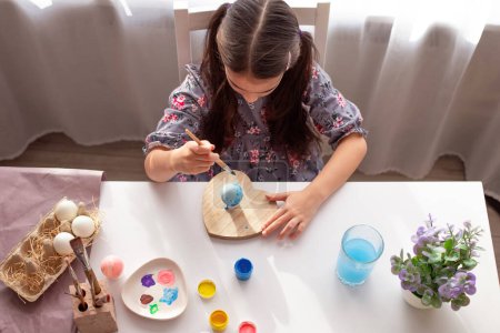 Photo for A little girl sits at a white table near the window, paints eggs with a brush. - Royalty Free Image