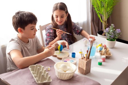 Photo for Smiling little girl and boy are sitting at a white table by the window, painting eggs for the Easter holiday - Royalty Free Image