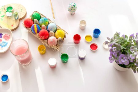 Photo for "On a white table, in daylight, there are brushes in a glass of water, multi-colored eggs in a tray, jars of paint." - Royalty Free Image