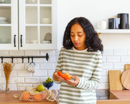 Téléchargez les photos : "Femme afro-américaine vérifiant agrumes orange pour la détérioration dans la cuisine après l'épicerie" - en image libre de droit