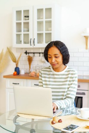 Téléchargez les photos : "Femme afro-américaine axée sur le travail à distance tout en étant assis dans la cuisine et en utilisant un ordinateur portable" - en image libre de droit