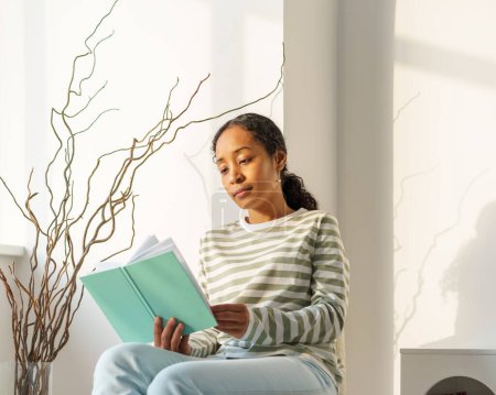 Téléchargez les photos : "Femme afro-américaine lisant paisiblement un livre dans le salon. Profiter du moment. Style de vie lent" - en image libre de droit