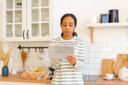 Téléchargez les photos : "Femme afro-américaine à la recherche de recette sur tablette pc avant de cuisiner le repas dans la cuisine" - en image libre de droit