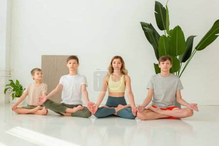 Téléchargez les photos : Famille sportive, une femme mince, un garçon et deux adolescents, assis en position lotus dans une pièce lumineuse - en image libre de droit