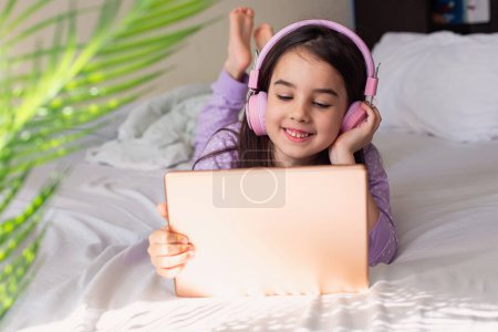 Photo for "Cute little girl in pink headphones, lying on the white bed, hold a pink digital tablet" - Royalty Free Image