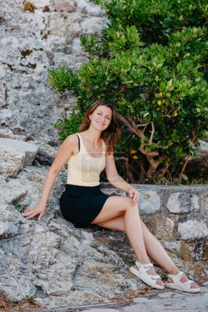 Téléchargez les photos : "Belle fille est assise sur le mur de pierre de la vieille ville de Budva. La fille est assise sur un parapet en pierre, Budva, Monténégro" - en image libre de droit