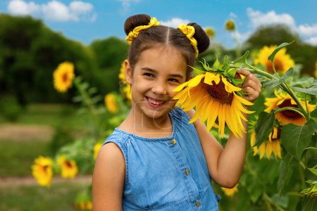 Photo for "A happy little girl in a blue dress stands next to the flowers of a decorative sunflower" - Royalty Free Image