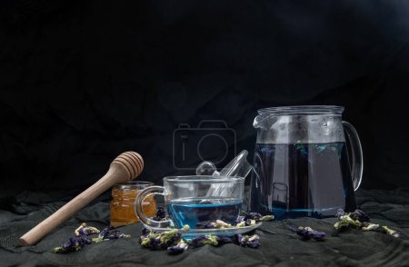 Téléchargez les photos : Thé au pois papillon ou ancan bleu biologique dans une théière et une tasse en verre servie avec du miel sur fond sombre - en image libre de droit