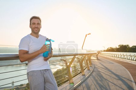 Foto de "Atleta seguro feliz, hombre caucásico de mediana edad en ropa deportiva sosteniendo una botella con agua, refrescándose después del entrenamiento y sonriendo mirando a la cámara mientras está de pie en un puente urbano al amanecer" - Imagen libre de derechos