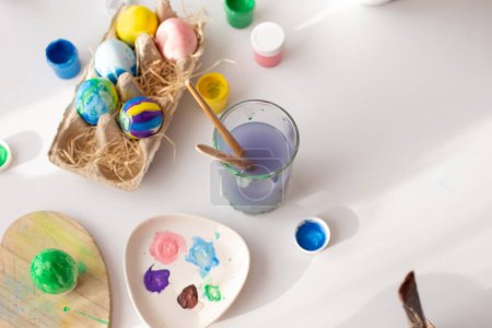 Photo for "On a white table, in daylight, there are brushes in a glass of water, multi-colored eggs in a tray, jars of paint" - Royalty Free Image