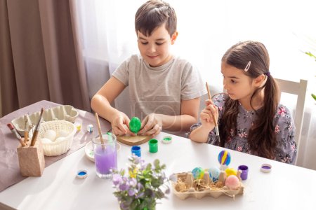 Photo for "A happy boy and little girl sits at a white table near the window, painting eggs" - Royalty Free Image