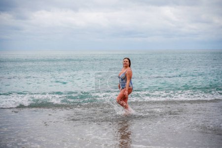 Téléchargez les photos : "Une femme dodue en maillot de bain pénètre dans l'eau pendant le surf. Seul sur la plage, Ciel gris dans les nuages, baignade en hiver." - en image libre de droit
