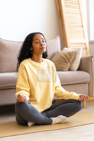 Téléchargez les photos : "Jeune femme afro-américaine assise dans une pose de yoga lotus sur tapis en méditant, verticale" - en image libre de droit