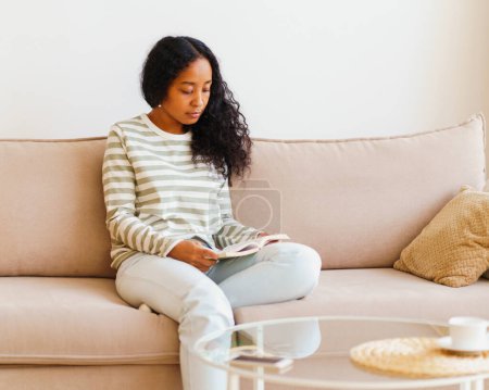 Téléchargez les photos : "Belle femme afro-américaine assise sur un canapé et lisant un livre. Passer du temps seul à la maison" - en image libre de droit