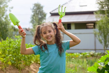 Téléchargez les photos : "Petite fille mignonne tenant de petits outils de jardinage, dansant dans le jardin" - en image libre de droit