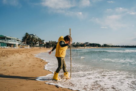 Téléchargez les photos : "Boy in yellow rubber boots playing with stick and sand at the beach. School kid touching water at autumn winter sea. Child having fun with waves at the shore. Spring Holiday vacation concept" - en image libre de droit
