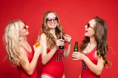 Photo for "Beautiful and happy female friends in bright red swimsuits with chilled drinks in their hands on a red background, studio." - Royalty Free Image