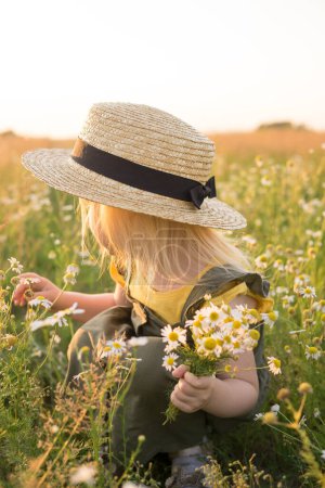 Téléchargez les photos : Petite fille blonde en chapeau de paille dans le champ avec un bouquet de marguerites - en image libre de droit
