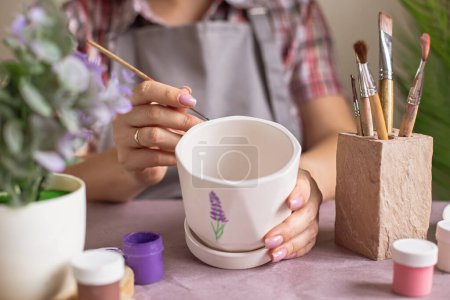 Téléchargez les photos : Les femmes en tablier gris peignent un pot en céramique de fleur blanche sur la table - en image libre de droit