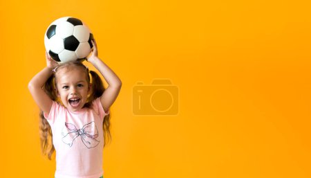Téléchargez les photos : Authentique mignon souriant préscolaire petite fille avec ballon de football classique noir et blanc regarder la caméra sur fond jaune. enfant jouer au football en t-shirt et short. Sport, championnat, concept d'équipe - en image libre de droit