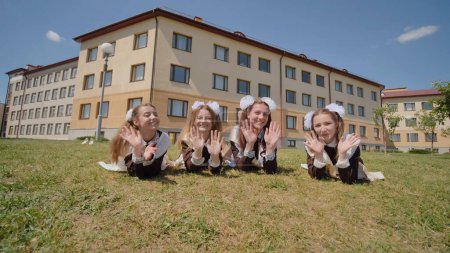 Photo for "Four schoolgirl graduates are lying on the grass against the backdrop of their school. Girls waving their hands and fleeing. Russian school." - Royalty Free Image