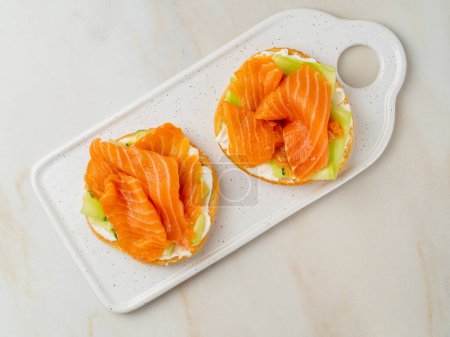 Foto de "Dos sándwiches abiertos con salmón, queso crema, rodajas de pepino sobre mesa de mármol blanco
" - Imagen libre de derechos