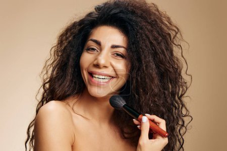 Photo for Enjoyed smiling adorable tanned sexy curly Latin makeup artist hold brush on cheek apply highlighter satisfied posing isolated on pastel beige background look at camera. Cosmetic offer concept - Royalty Free Image