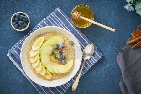 Photo for Oatmeal with bananas, blueberries, jam, honey, blue napkin on blue stone background - Royalty Free Image