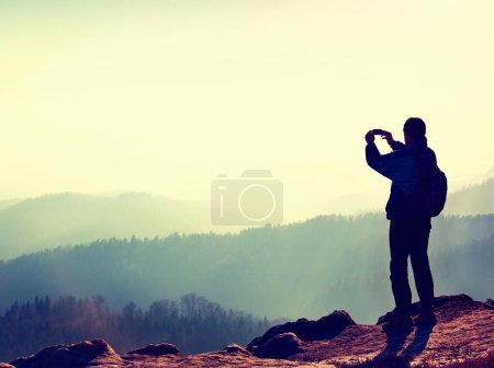 Photo for "Tourist takes photos with smart phone on peak of rock. Dreamy fogy landscape, spring orange pink misty sunrise in a beautiful valley below rocky mountains." - Royalty Free Image