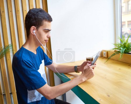 Photo for Young adult texting message on smart phone and listening to music on earphones in coffee shop - Royalty Free Image
