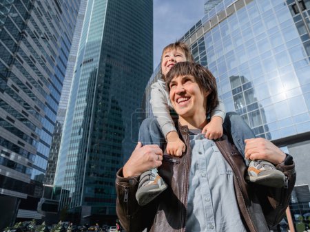 Foto de Little boy sits on father's shoulders among skyscrapers. Dad and son looks on glass walls of buildings. Future and modern technologies, life balance and family life in well keeps districts. - Imagen libre de derechos