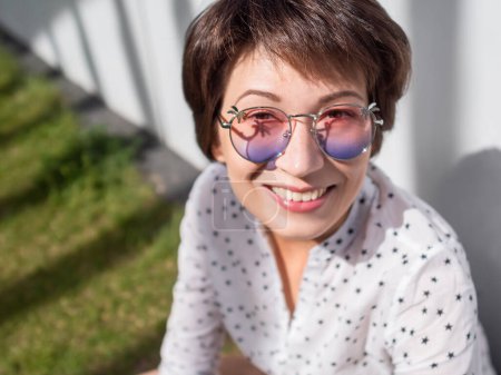 Téléchargez les photos : "Femme souriante dans des lunettes de soleil colorées a un repos sur la pelouse dans le parc urbain. La nature en ville. Détendez-vous à l'extérieur après le travail. Ambiances estivales." - en image libre de droit