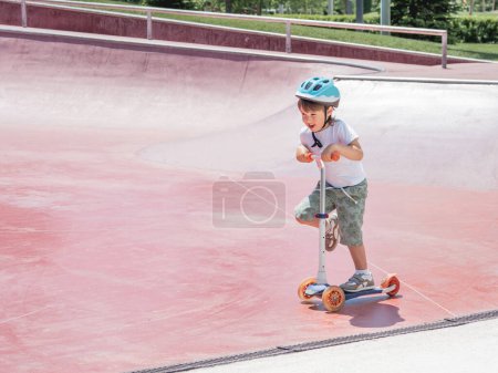 Foto de "Los paseos de niños pequeños patean scooter en el parque de patinaje. Estructuras especiales de cuenco de hormigón en parque urbano. Entrenamiento para patinar en verano." - Imagen libre de derechos