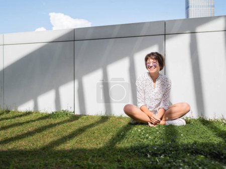 Téléchargez les photos : "Femme souriante dans des lunettes de soleil colorées a un repos sur la pelouse dans le parc urbain. La nature en ville. Détendez-vous à l'extérieur après le travail. Ambiances estivales." - en image libre de droit