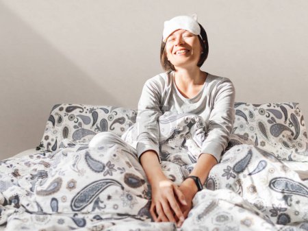 Photo for "Smiling woman in grey pajama and sleeping mask. She is just woke up and sit in bed. Early morning in cozy home." - Royalty Free Image