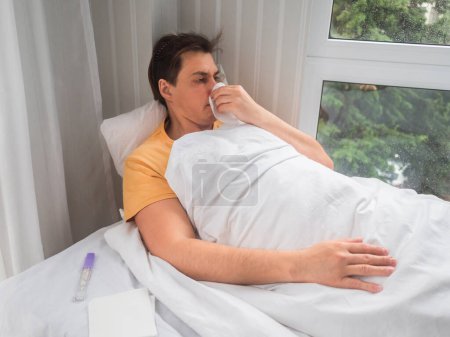 Téléchargez les photos : "Le malade est couché au lit avec un thermomètre. L'homme se mouche. Invalidité temporaire en cas de symptômes de la maladie." - en image libre de droit