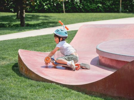 Foto de El niño se cayó del patinete scooter mientras montaba en el parque de skate. Estructuras especiales de cuenco de hormigón en parque urbano. Entrenamiento para patinar en verano. - Imagen libre de derechos