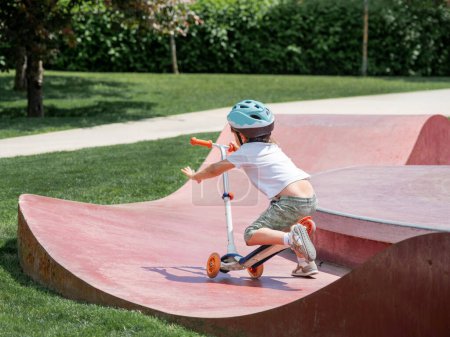 Photo for Little boy fell off kick scooter while riding in skate park. Special concrete bowl structures in urban park. Training to skate at summer. - Royalty Free Image
