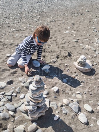 Téléchargez les photos : Petit garçon construit une pyramide avec des pierres et du bois flotté sur la plage de galets. Vacances au bord de la mer. Explorer la nature dans l'enfance. - en image libre de droit