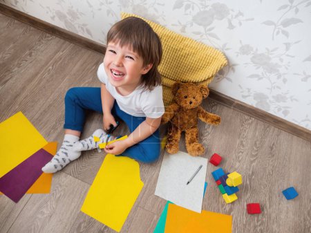 Photo for Toddler boy learns to cut colored paper with scissors. Kid sits on floor in kids room with toy blocks and teddy bear. Educational classes for children. Developing feeling sensations and fine motor skills at home. - Royalty Free Image